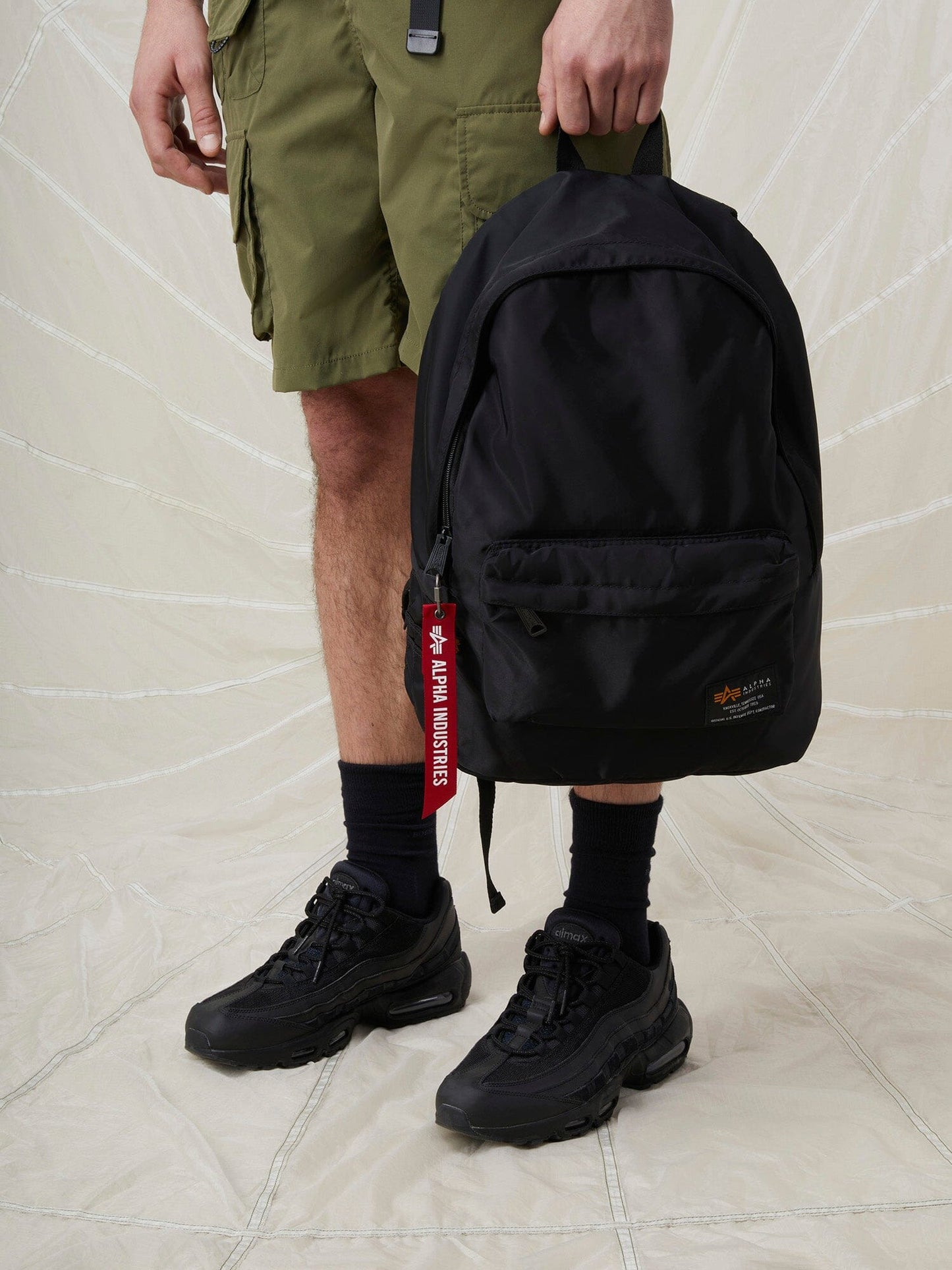 A person carrying a Crew Backpack by Alpha Industries, a sleek black backpack with ultra lightweight design and adjustable nylon straps.