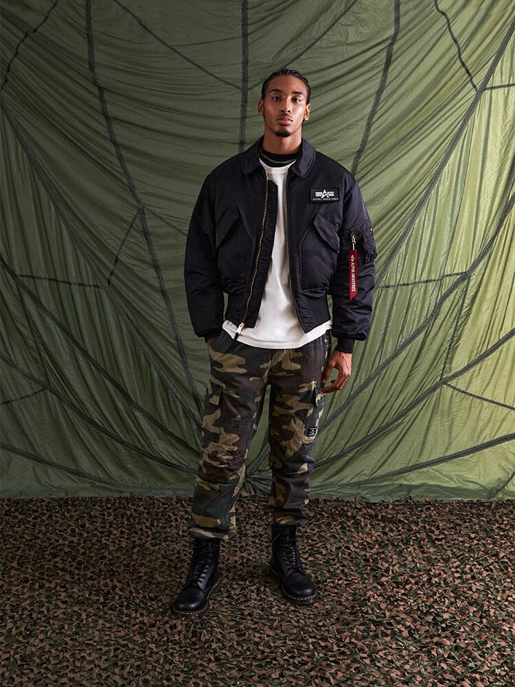 Alpha Industries Cwu 45/P Bomber Jacket in green, white t-shirt, camouflage pants, black boots.