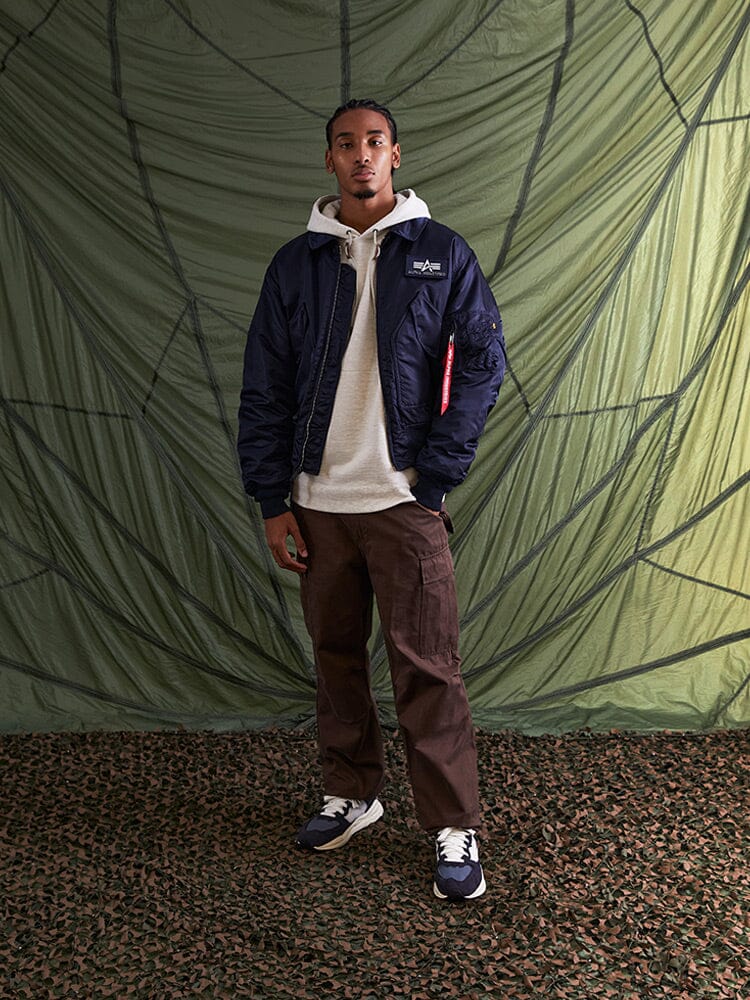 Alpha Industries Cwu 45/P Bomber Jacket, beige hoodie, brown cargo pants, sneakers; camouflage floor not shown.