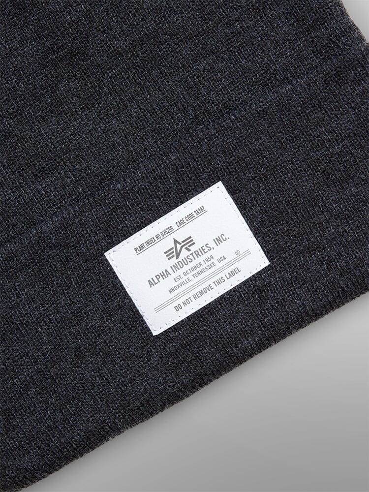 Close-up of the Essential Beanie in black featuring a white paper label from Alpha Industries, Inc., showcasing branding and care instructions.