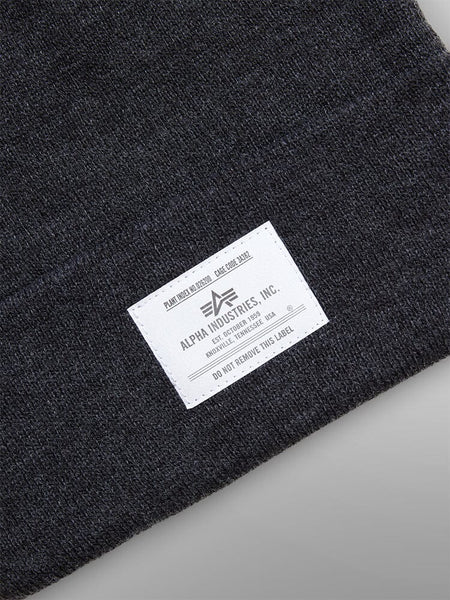Close-up of the Essential Beanie in black featuring a white paper label from Alpha Industries, Inc., showcasing branding and care instructions.