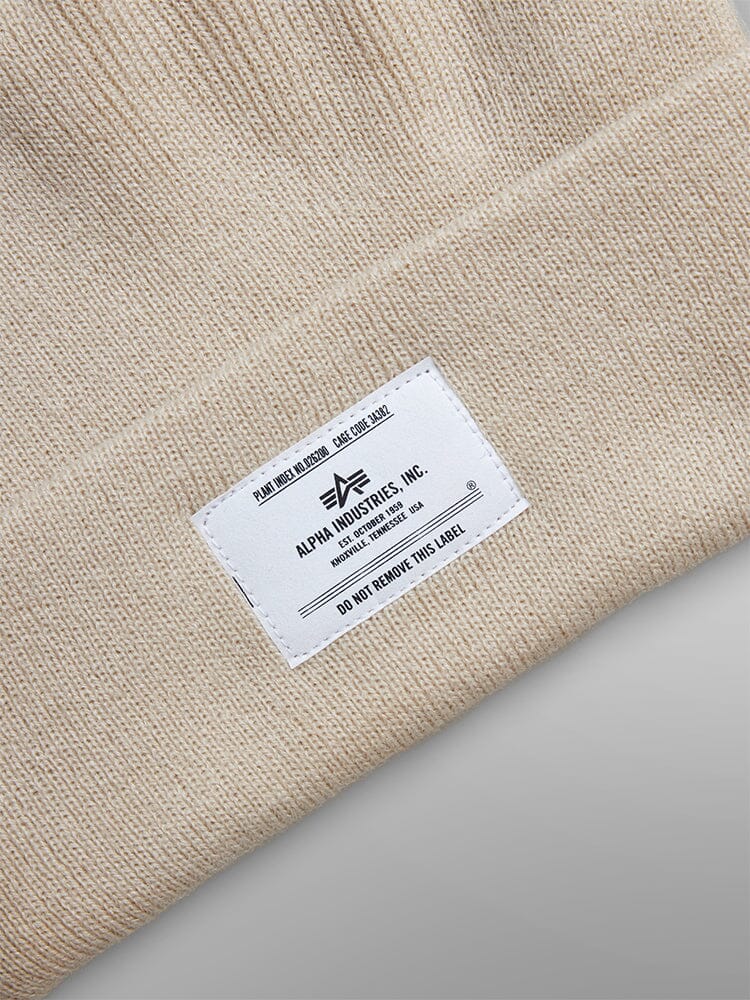 Beige knit beanie from Alpha Industries, Inc. with a white paper label featuring care instructions and the iconic logo, known as the Essential Beanie.