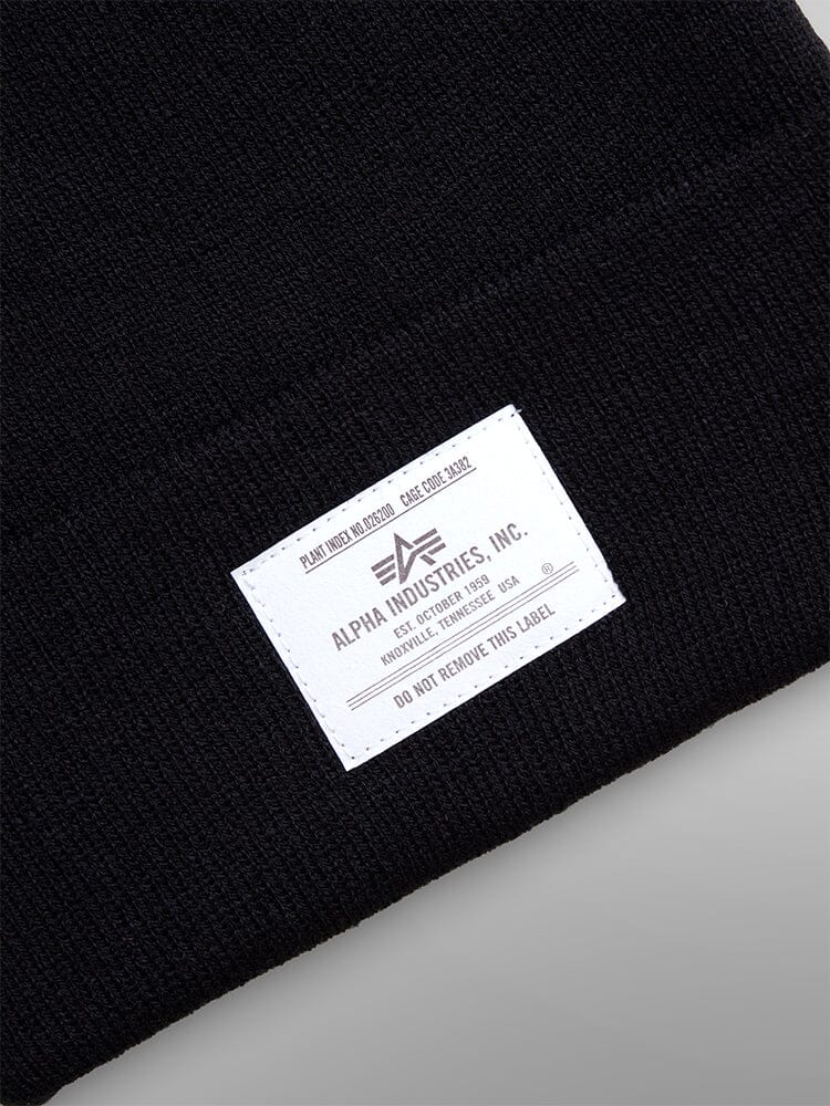 Close-up of the Essential Beanie in black featuring an Alpha Industries, Inc. paper label with care instructions and manufacturing details.