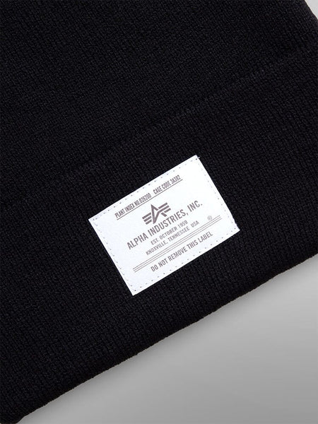 Close-up of the Essential Beanie in black featuring an Alpha Industries, Inc. paper label with care instructions and manufacturing details.