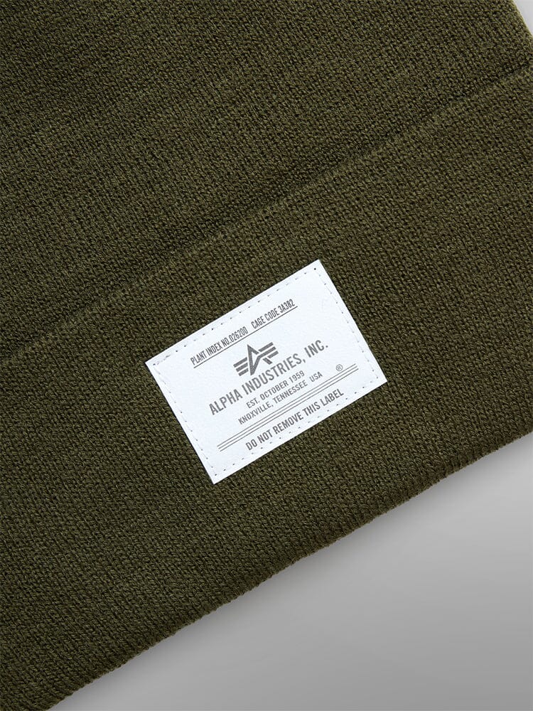 The Essential Beanie by Alpha Industries is an olive green knit cap adorned with a white label that reads "Remove Before Flight" and includes care instructions.
