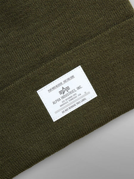 The Essential Beanie by Alpha Industries is an olive green knit cap adorned with a white label that reads "Remove Before Flight" and includes care instructions.