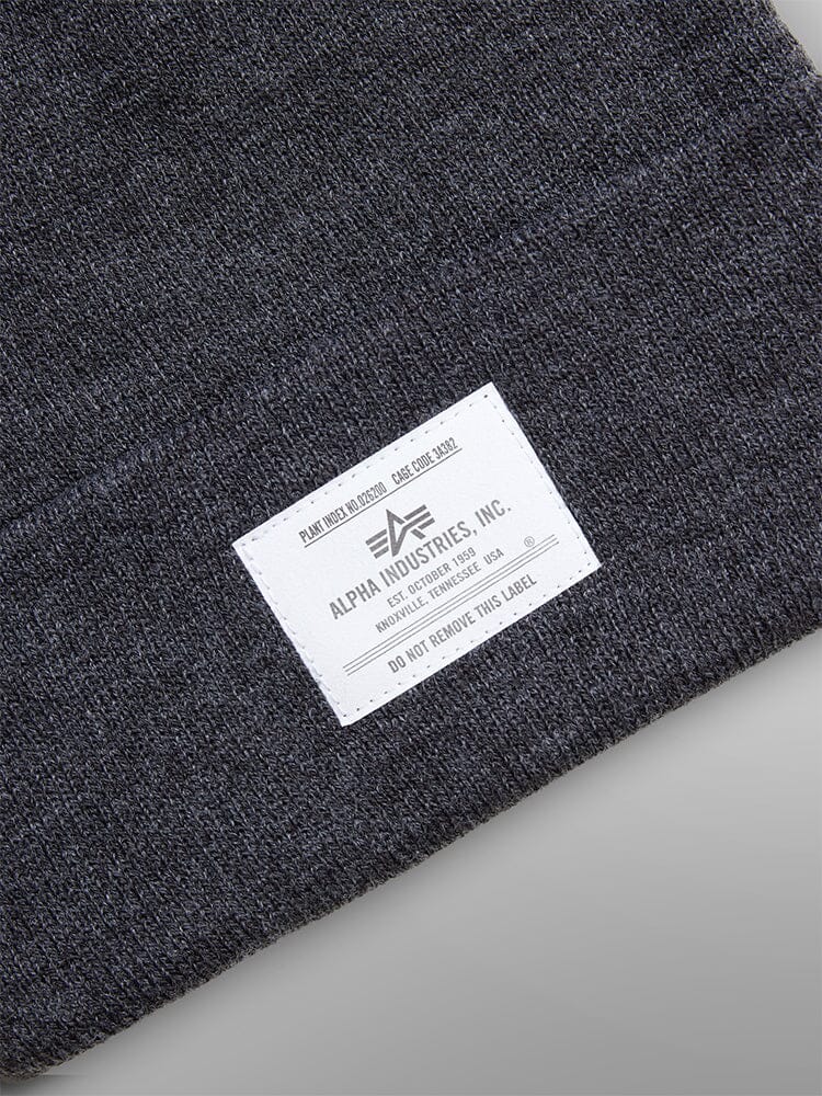 Close-up of a Dark Gray Essential Beanie by Alpha Industries, Inc., showcasing a white label with manufacturing details and care instructions.