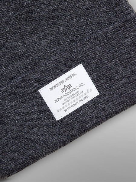 Close-up of a Dark Gray Essential Beanie by Alpha Industries, Inc., showcasing a white label with manufacturing details and care instructions.