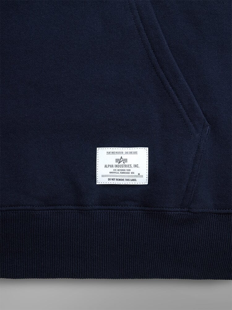 Close-up of the Essential Hoodie in dark blue, by Alpha Industries. Showcasing a white label by the hem and a convenient pocket.