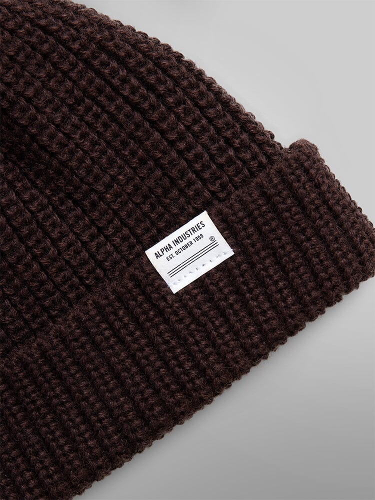 Close-up of the Essential Watch Cap, a brown ribbed wool-blend beanie reminiscent of a classic watch cap, featuring a white label with the Alpha Industries logo.