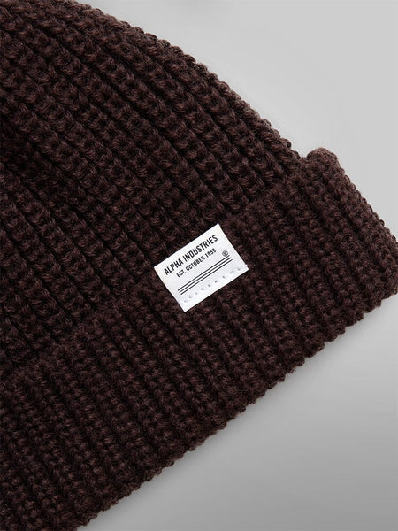 Close-up of the Essential Watch Cap, a brown ribbed wool-blend beanie reminiscent of a classic watch cap, featuring a white label with the Alpha Industries logo.