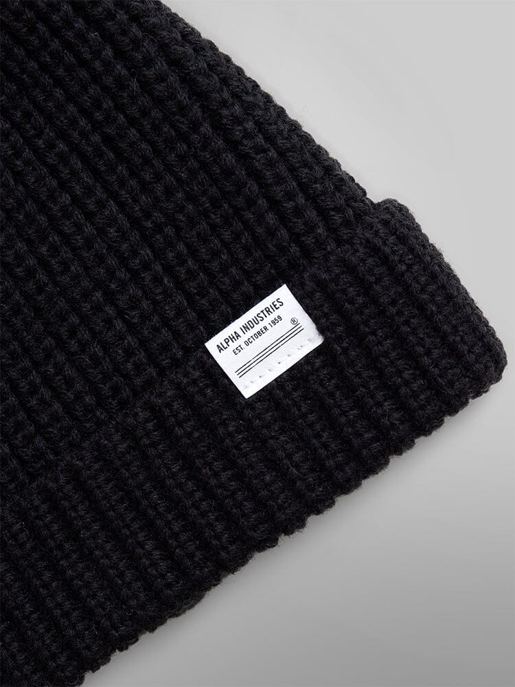 Close-up of an Essential Watch Cap, a black wool-blend knit beanie featuring a white Alpha Industries label sewn on the folded edge, reminiscent of classic watch caps worn throughout military history.