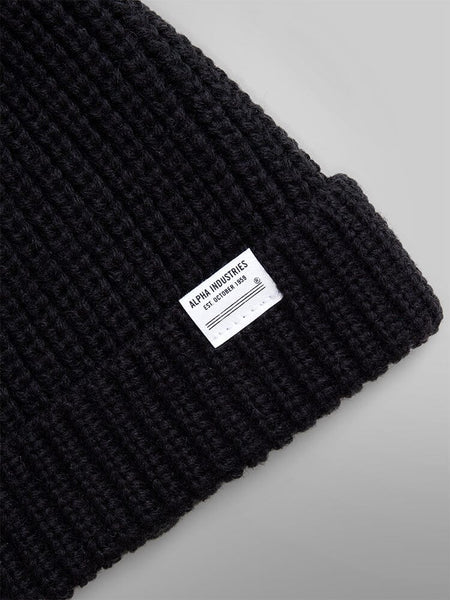 Close-up of an Essential Watch Cap, a black wool-blend knit beanie featuring a white Alpha Industries label sewn on the folded edge, reminiscent of classic watch caps worn throughout military history.