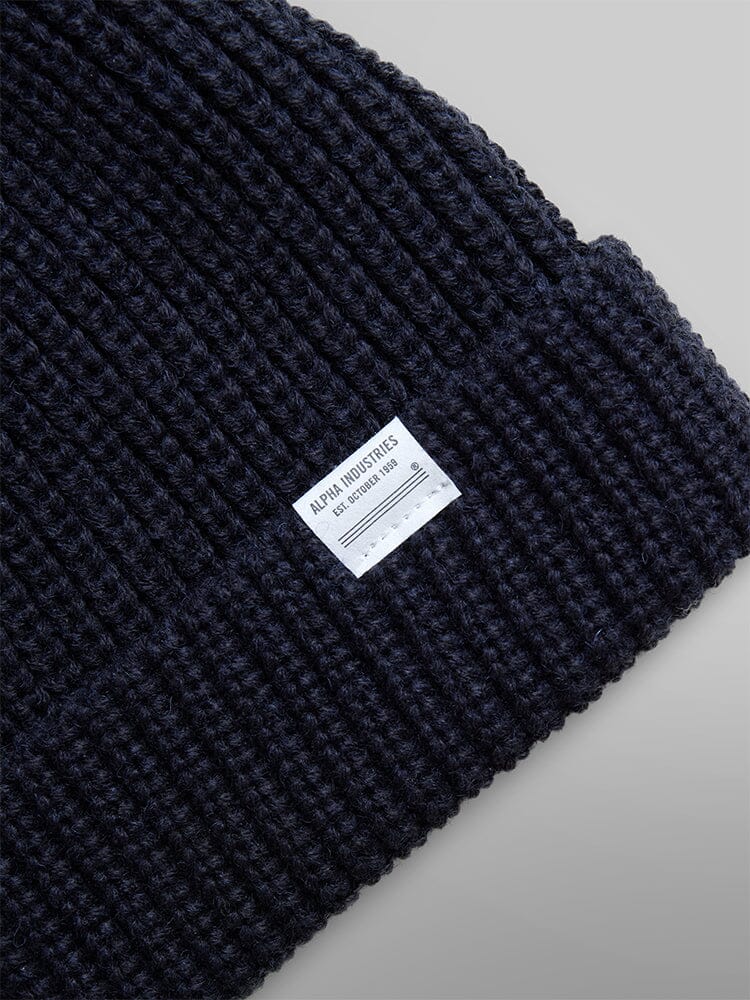 The Essential Watch Cap by Alpha Industries is a black ribbed knit beanie made from a wool-blend material, featuring a white label on the fold..