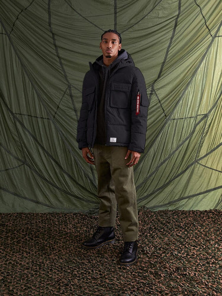 A man wearing the Hooded Puffer Jacket by Alpha Industries, Inc. and olive green pants stands on a textured surface.
