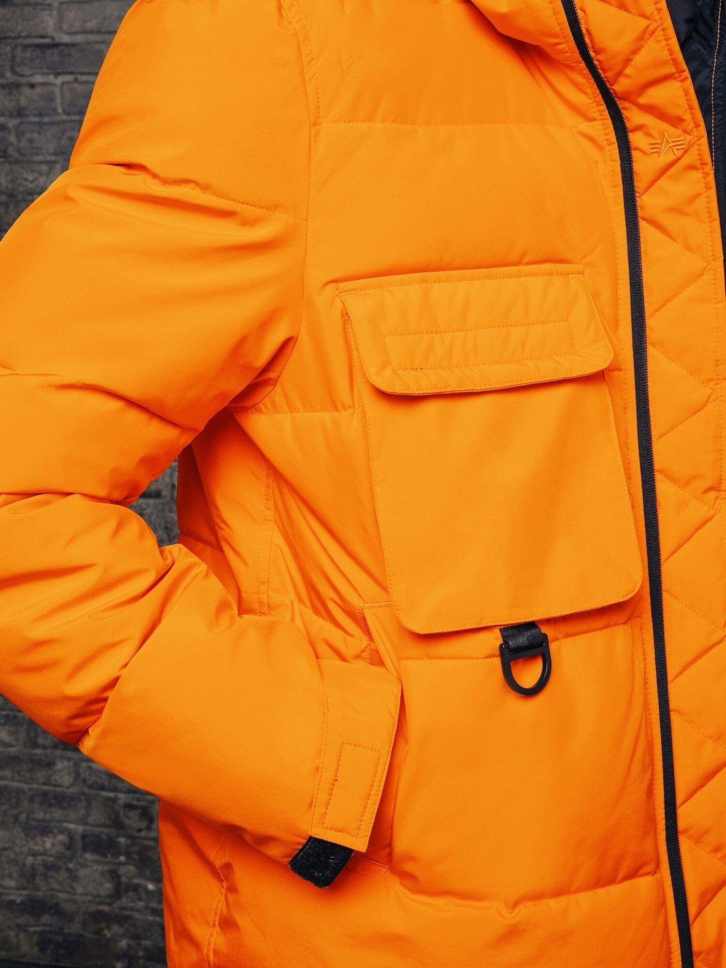 A close-up of an individual sporting the Alpha Industries, Inc. Hooded Puffer Jacket in vibrant orange showcases a sleek black zipper and detailing. This stylish piece includes a large front pocket with a D-ring. The nylon design features an insulated hood to ensure warmth during chilly weather conditions.