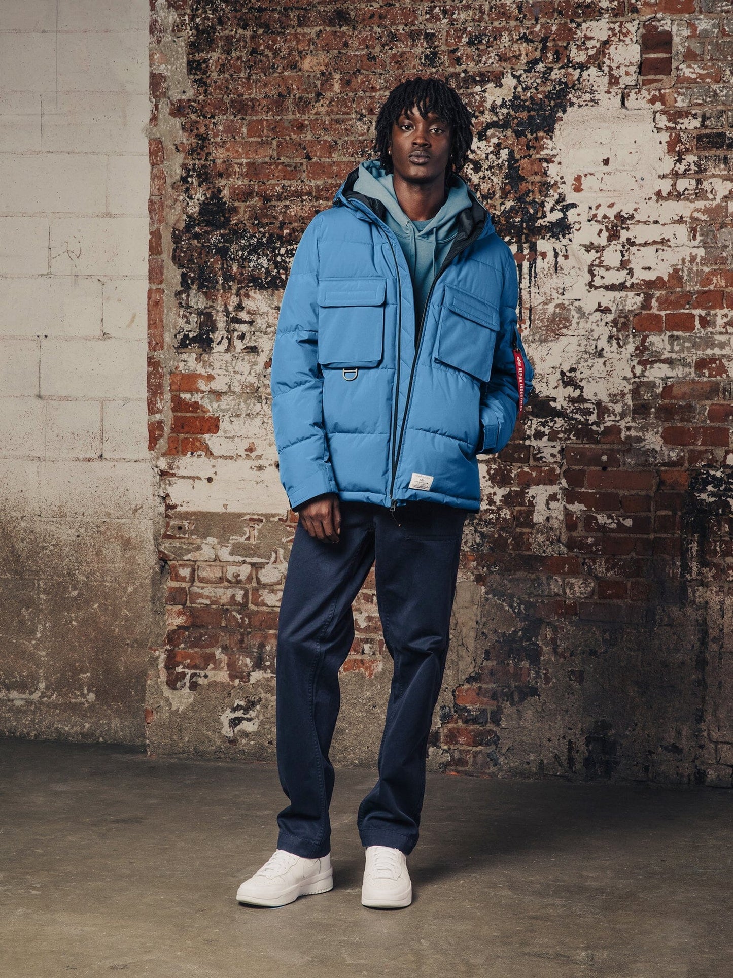 A person wearing a Hooded Puffer Jacket by Alpha Industries, Inc., along with blue jeans and white sneakers.
