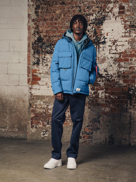 A person wearing a Hooded Puffer Jacket by Alpha Industries, Inc., along with blue jeans and white sneakers.