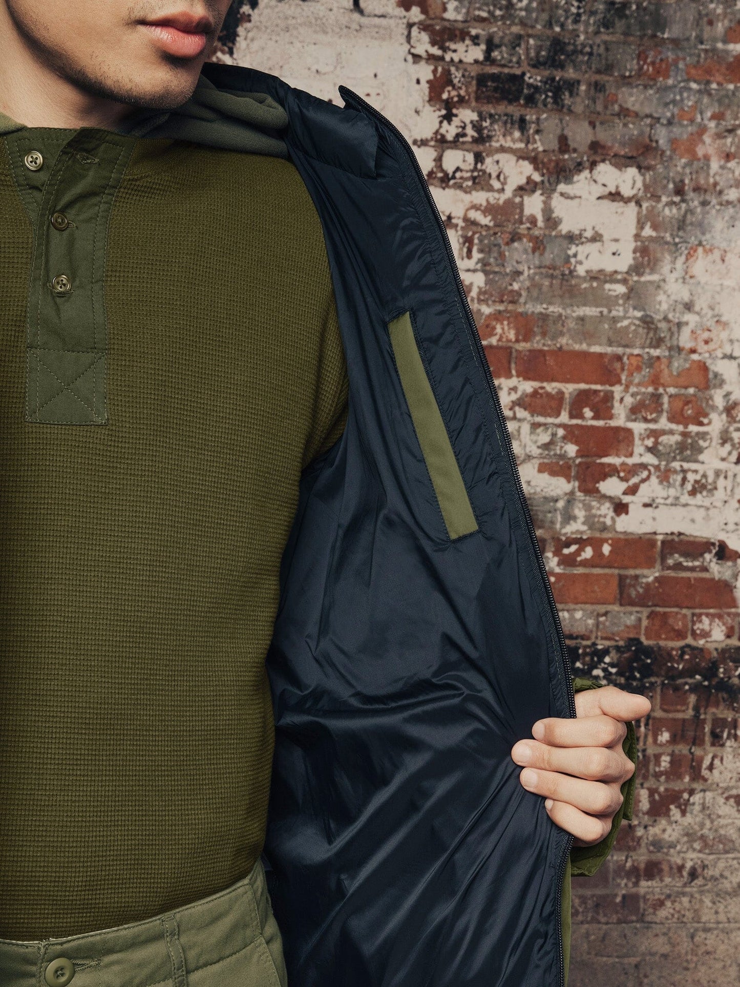 A person unzips an Alpha Industries Hooded Puffer Jacket in a dark color, revealing a green shirt underneath.