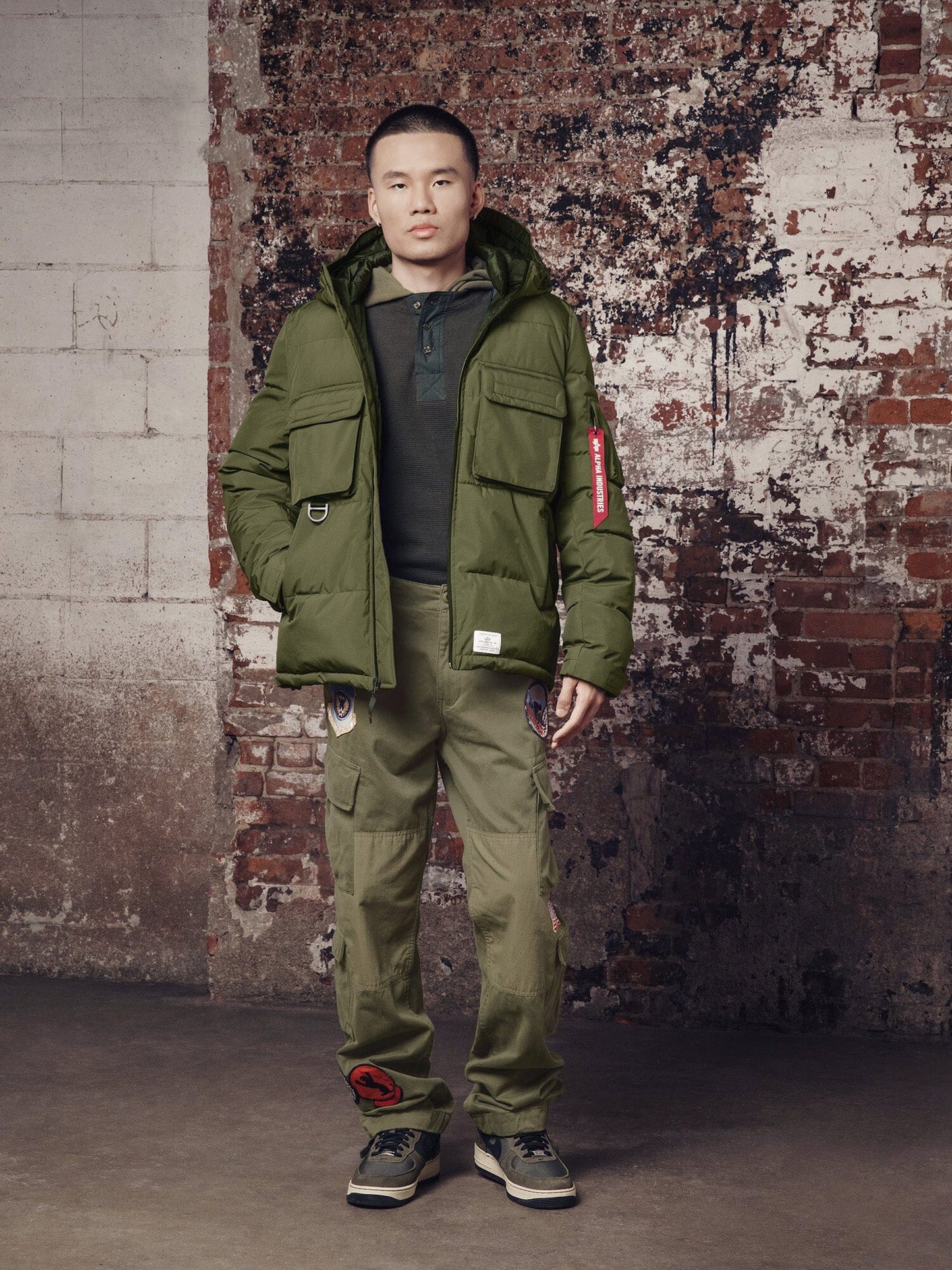 A person stands sporting the Hooded Puffer Jacket by Alpha Industries, Inc., paired with matching green cargo pants with patches and complemented by gray sneakers.