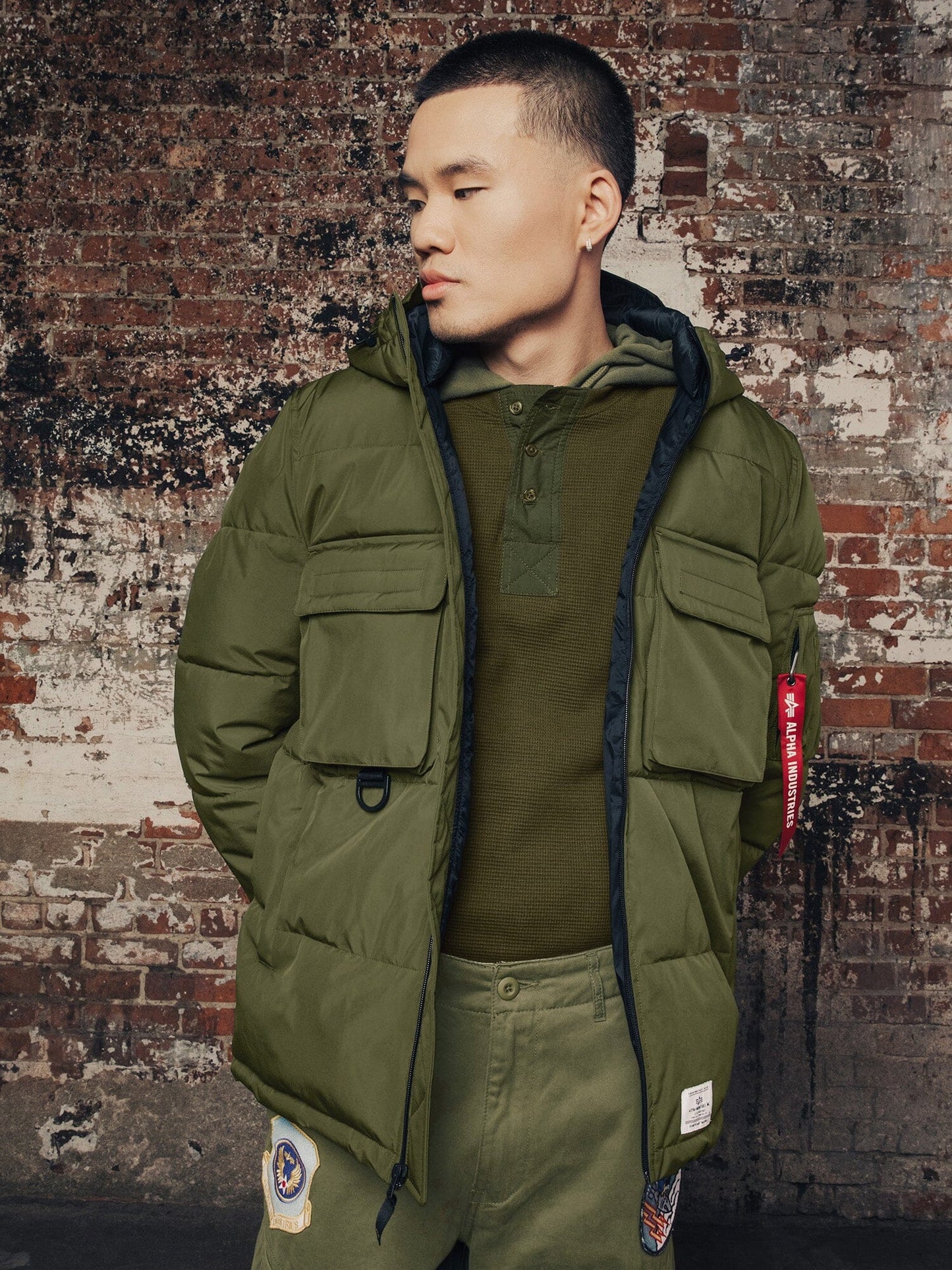 A person poses wearing the Hooded Puffer Jacket from Alpha Industries, Inc. This green nylon jacket with an insulated hood is perfectly complemented by a matching green shirt and patched green pants.