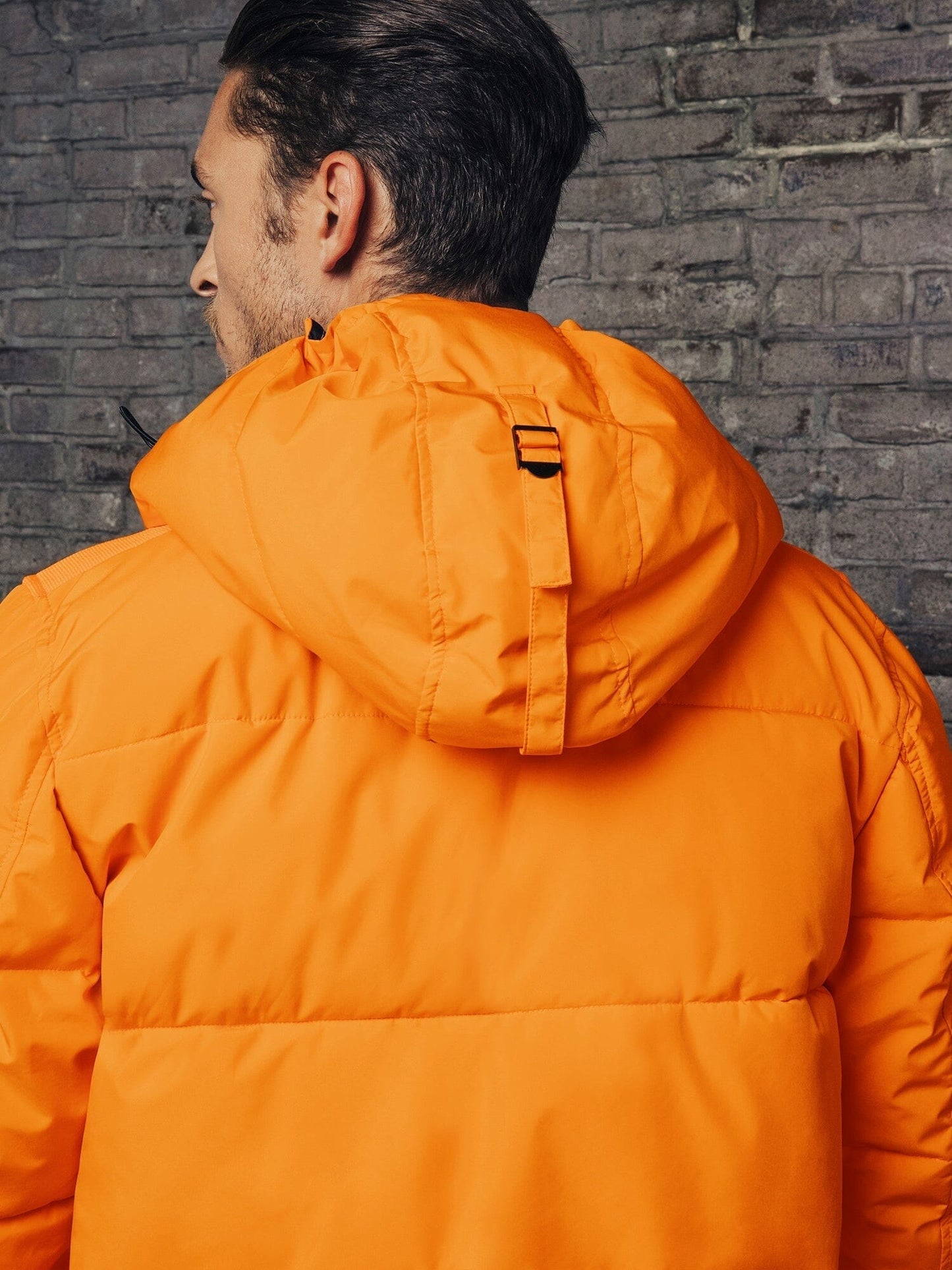 A person wearing an Alpha Industries Hooded Puffer Jacket in orange, featuring an insulated hood, is facing a brick wall.