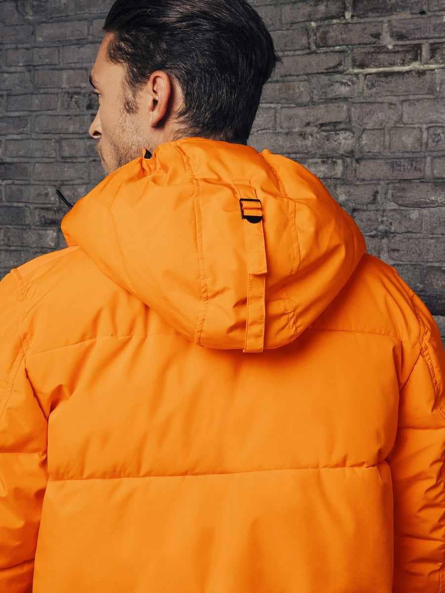 A person wearing an Alpha Industries Hooded Puffer Jacket in orange, featuring an insulated hood, is facing a brick wall.