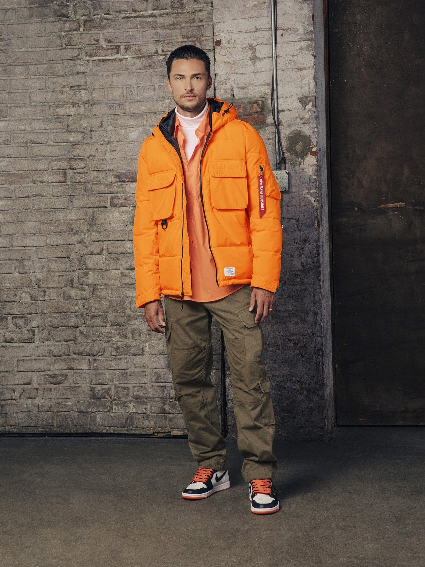 A person wearing an Alpha Industries, Inc. Hooded Puffer Jacket in orange, paired with an orange shirt, khaki pants, and sneakers, stands with a neutral expression.