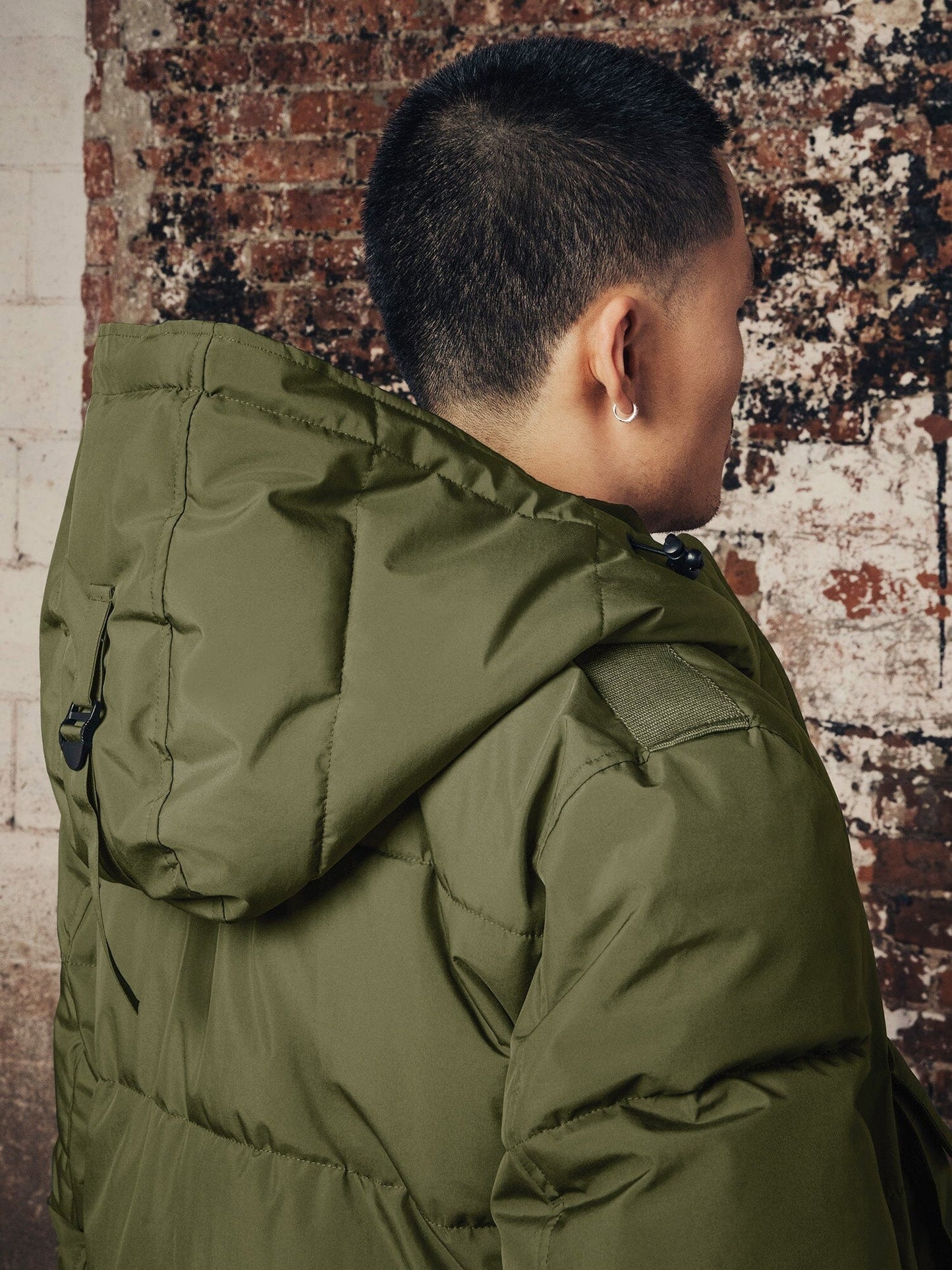A person stands with their back to the camera, comfortably clad in an olive green Hooded Puffer Jacket by Alpha Industries, Inc.