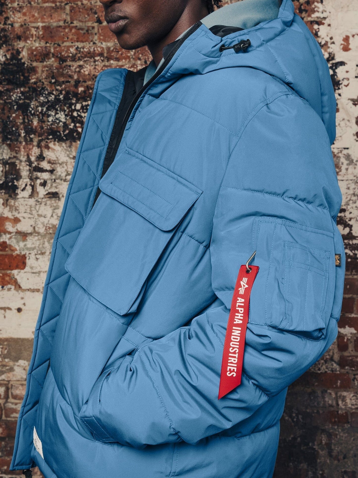 A person wearing the Hooded Puffer Jacket by Alpha Industries, Inc., featuring blue nylon fabric, multiple pockets, an insulated hood, and a red Remove Before Flight tag.