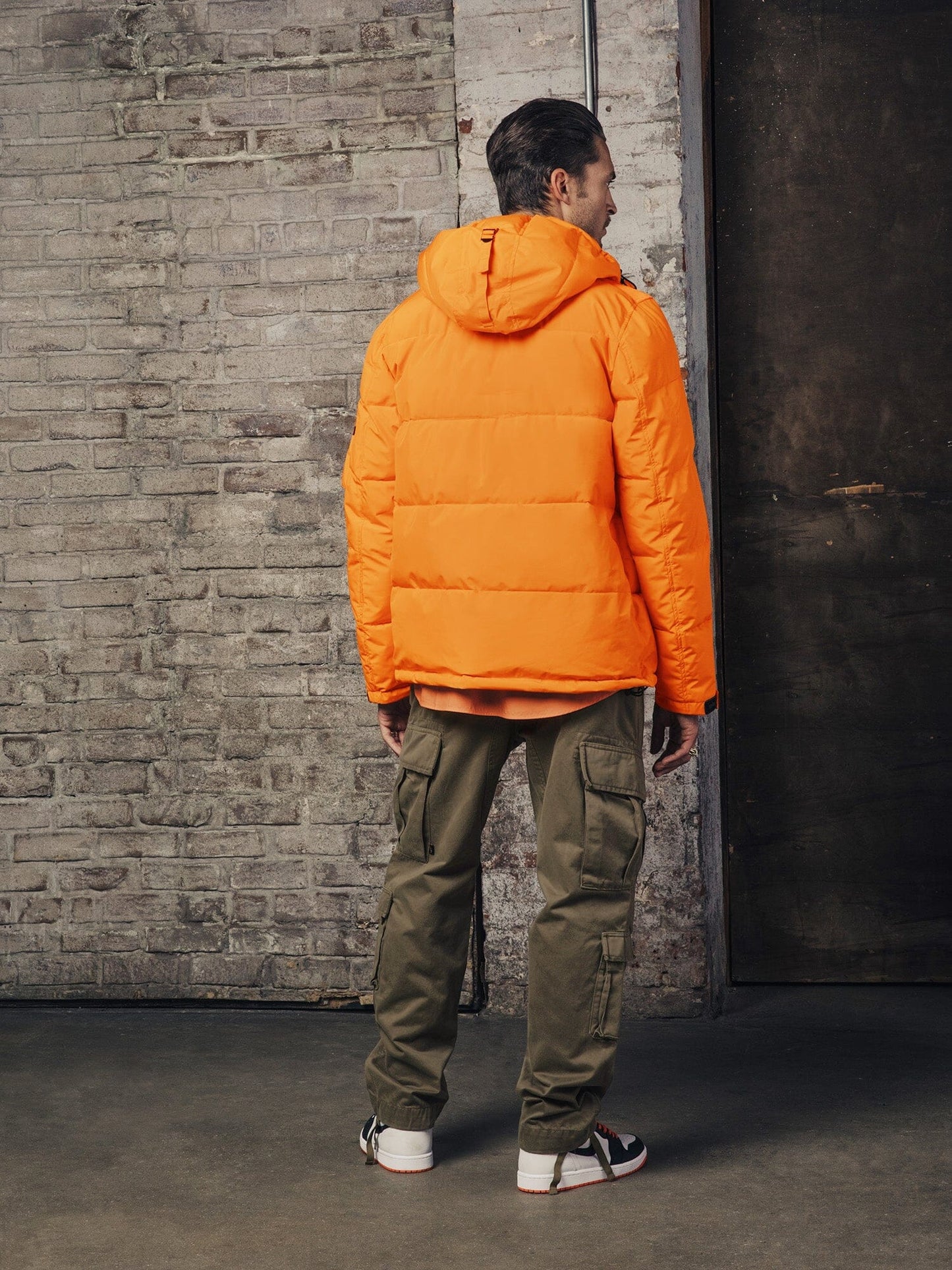 An individual wearing the Hooded Puffer Jacket by Alpha Industries, Inc., in an orange nylon material with an insulated hood, and pairing it with khaki cargo pants is standing facing a brick wall.