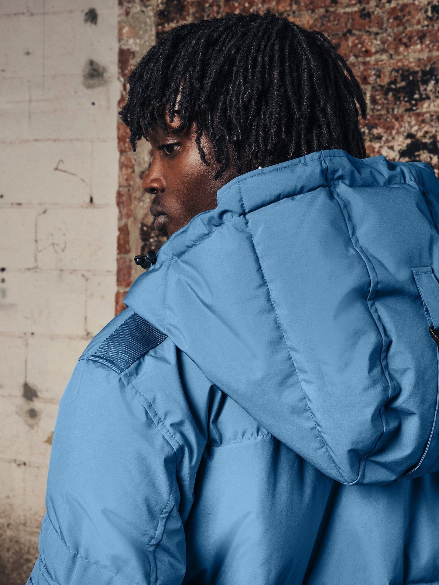 A person with dreadlocks wearing the Hooded Puffer Jacket by Alpha Industries, Inc. stands against a brick and concrete wall.