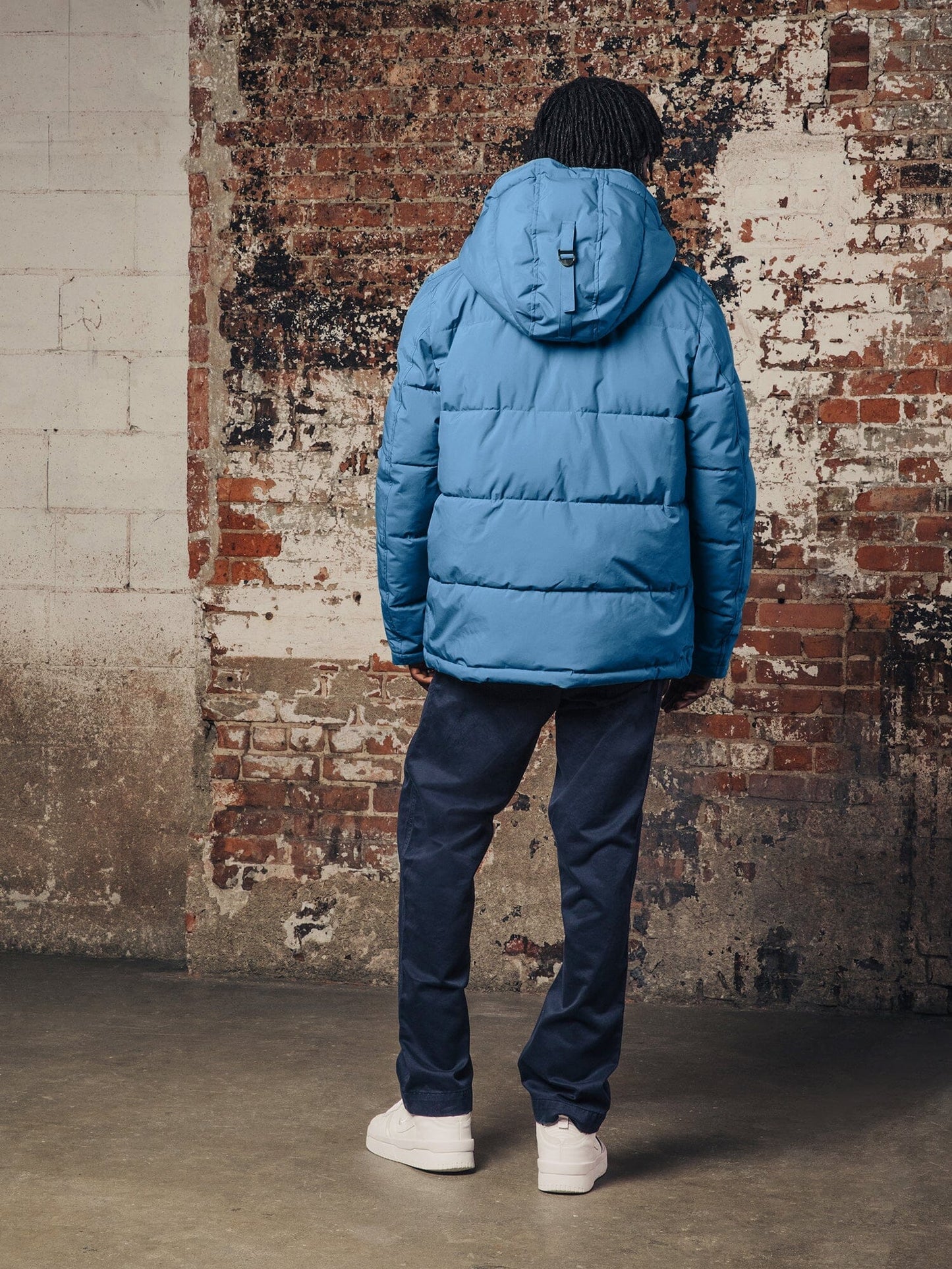 A person wearing the Hooded Puffer Jacket by Alpha Industries, Inc., in blue nylon with an insulated hood, paired with dark pants, stands facing a textured brick and concrete wall.