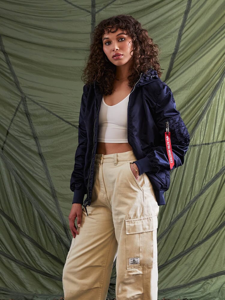 A woman with curly hair stands wearing the L-2B Hooded Gen II Bomber Jacket by Alpha Industries, Inc., paired with a white top and beige cargo pants.