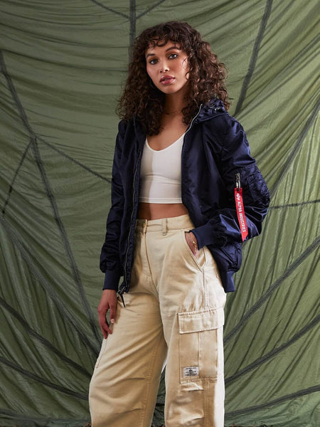 A woman with curly hair stands wearing the L-2B Hooded Gen II Bomber Jacket by Alpha Industries, Inc., paired with a white top and beige cargo pants.