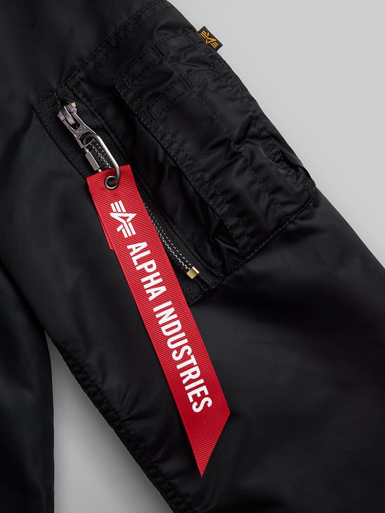 Close-up of a black nylon sleeve from the L-2B Hooded Gen II Bomber Jacket by Alpha Industries, Inc., displaying a zippered pocket and a red tag with the brand's text and logo, highlighting the sleek design of this water-resistant flight jacket.