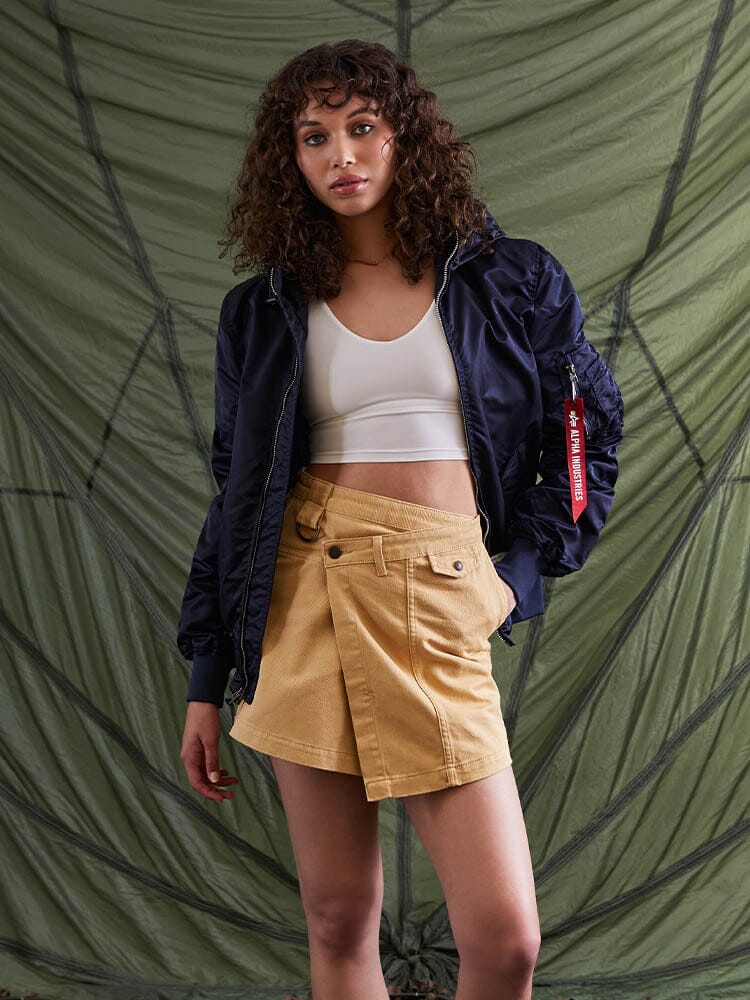 A person with curly hair, dressed in an Alpha Industries L-2B Hooded Gen II Bomber Jacket, a white crop top, and a tan skirt.