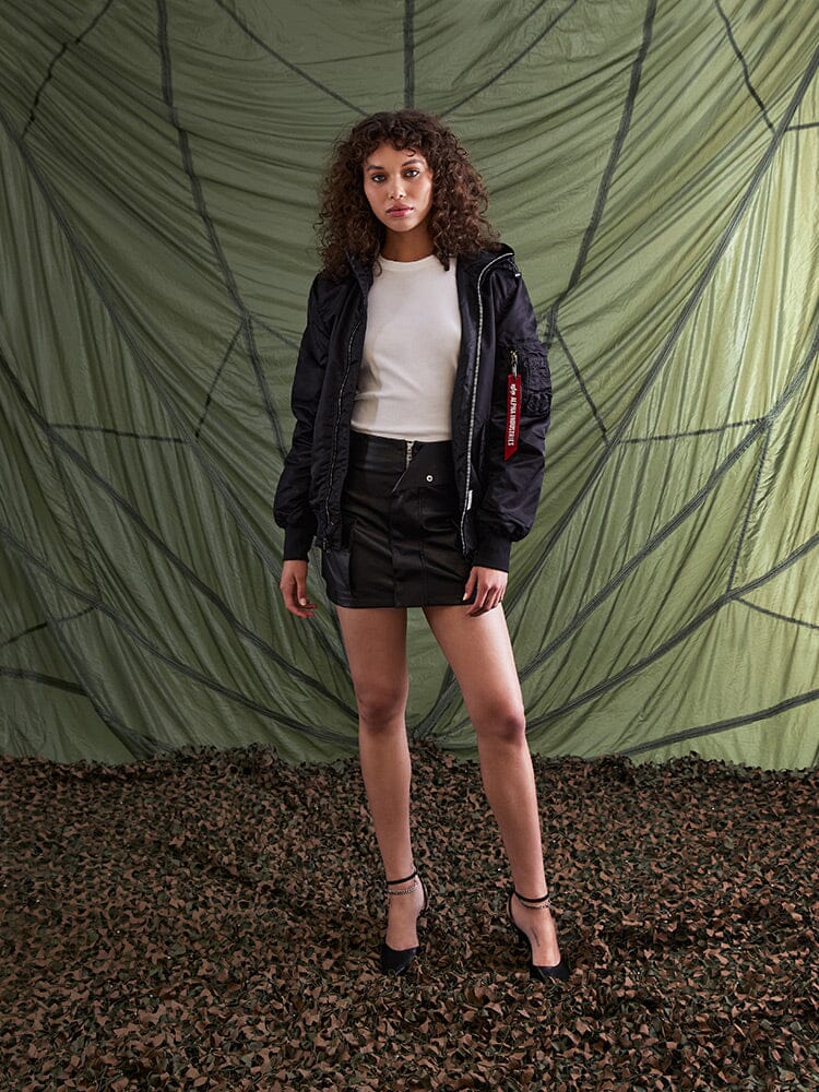 A person stands wearing the L-2B Hooded Gen II Bomber Jacket by Alpha Industries, Inc., paired with a white shirt, black skirt, and heels.