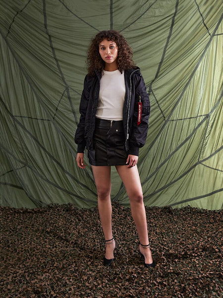 A person stands wearing the L-2B Hooded Gen II Bomber Jacket by Alpha Industries, Inc., paired with a white shirt, black skirt, and heels.