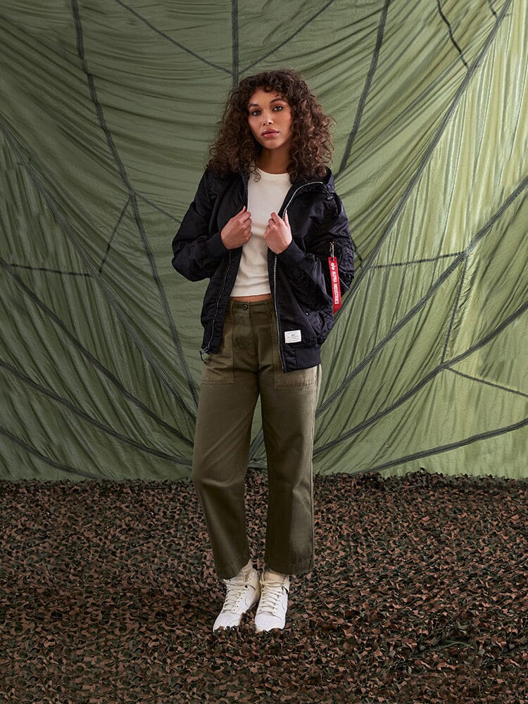 A person with curly hair stands wearing an Alpha Industries L-2B Hooded Gen II Bomber Jacket, a white shirt, olive pants, and white shoes.