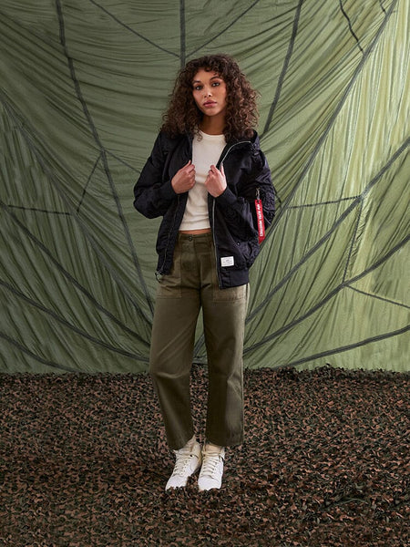 A person with curly hair stands wearing an Alpha Industries L-2B Hooded Gen II Bomber Jacket, a white shirt, olive pants, and white shoes.