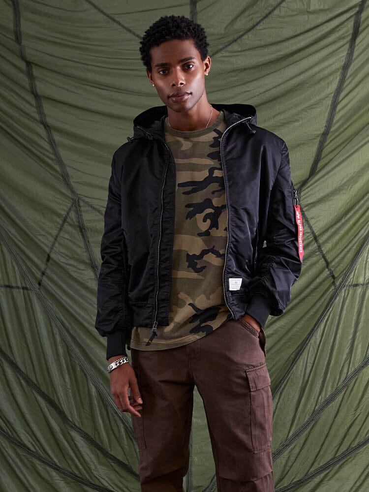 A person wearing an Alpha Industries L-2B Hooded Gen II Bomber Jacket and a camouflage shirt.