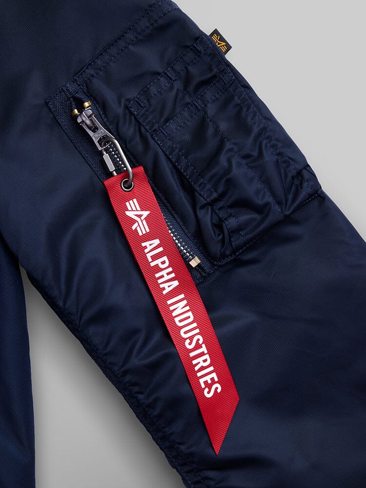 A close-up of a navy pocket on the L-2B Hooded Gen II bomber jacket, crafted from water-resistant nylon, showcases a silver zipper with a red fabric tag labeled Alpha Industries, Inc. attached to the zipper pull.
