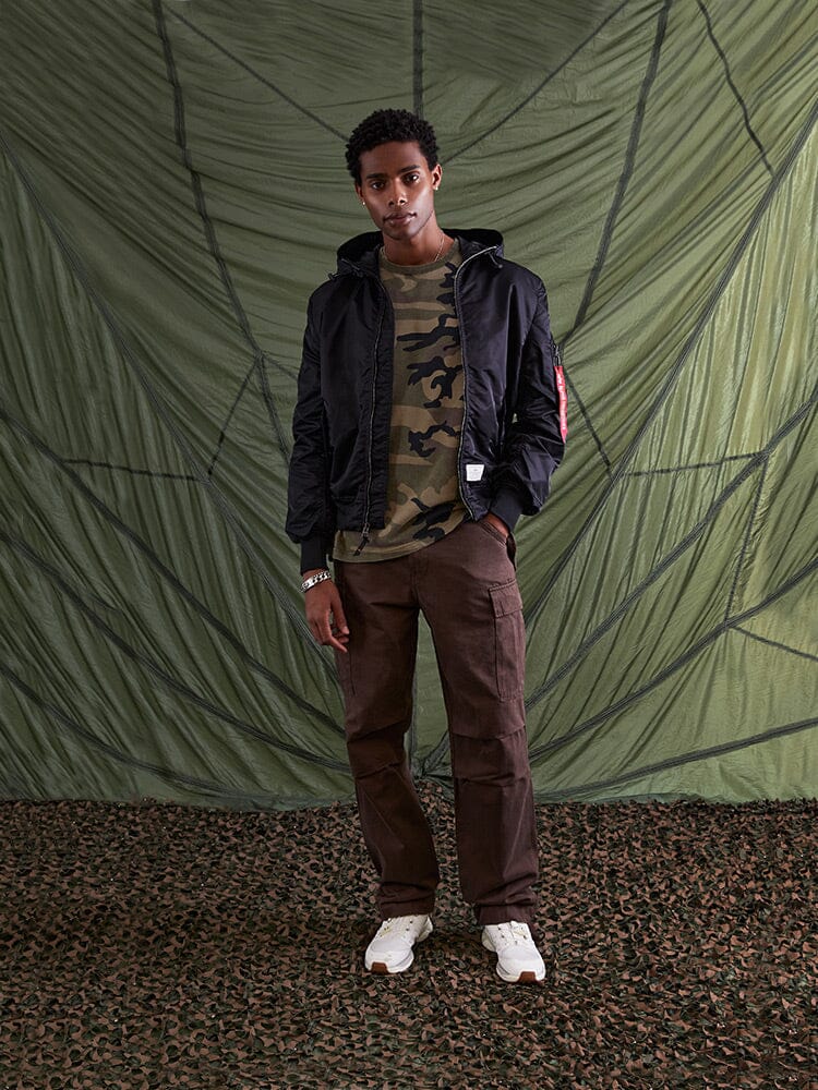 The individual poses dressed in the L-2B Hooded Gen II Bomber Jacket by Alpha Industries, Inc., over a camouflage shirt, paired with brown pants and white sneakers.