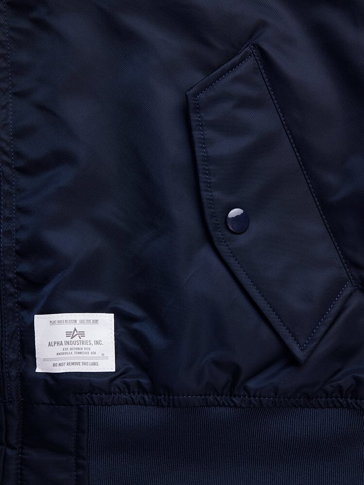 Close-up of a navy Alpha Industries L-2B Hooded Gen II Bomber Jacket featuring a large pocket with a snap button and a white manufacturer's label stitched at the bottom.