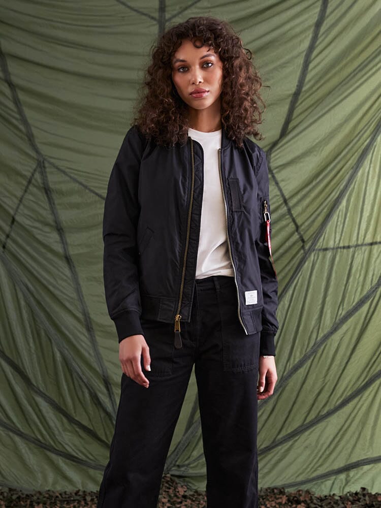 A person with curly hair wearing an Alpha Industries L-2B Skymaster Gen II Bomber Jacket W and a white shirt stands.