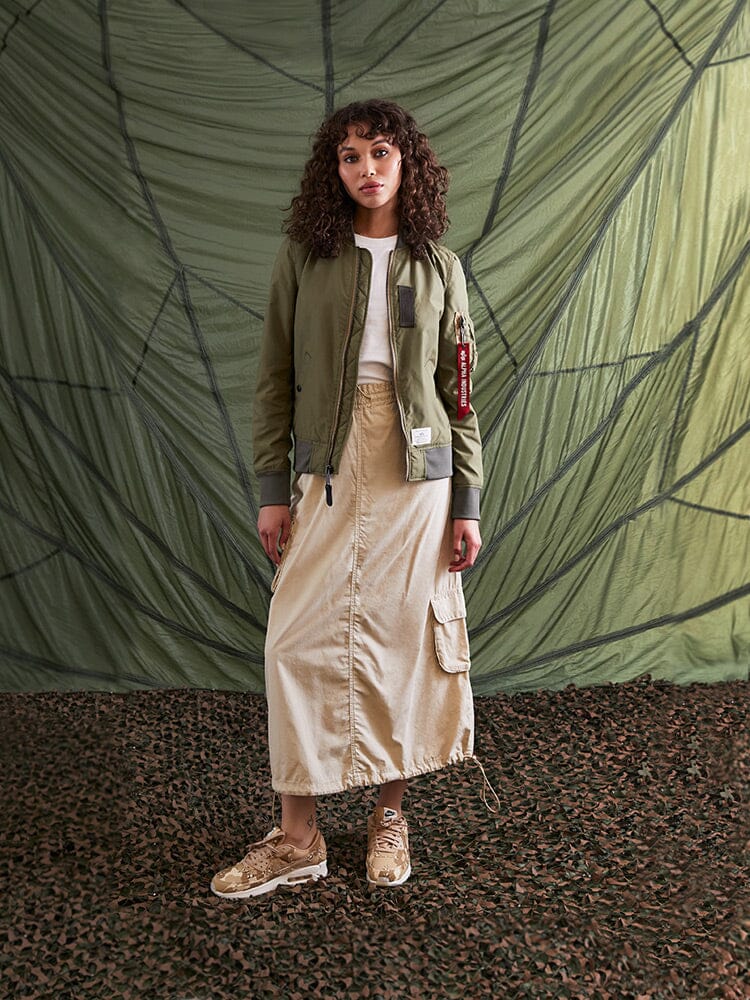 A person stands donned in the L-2B Skymaster Gen II Bomber Jacket by Alpha Industries, Inc., paired with a beige skirt and sneakers.