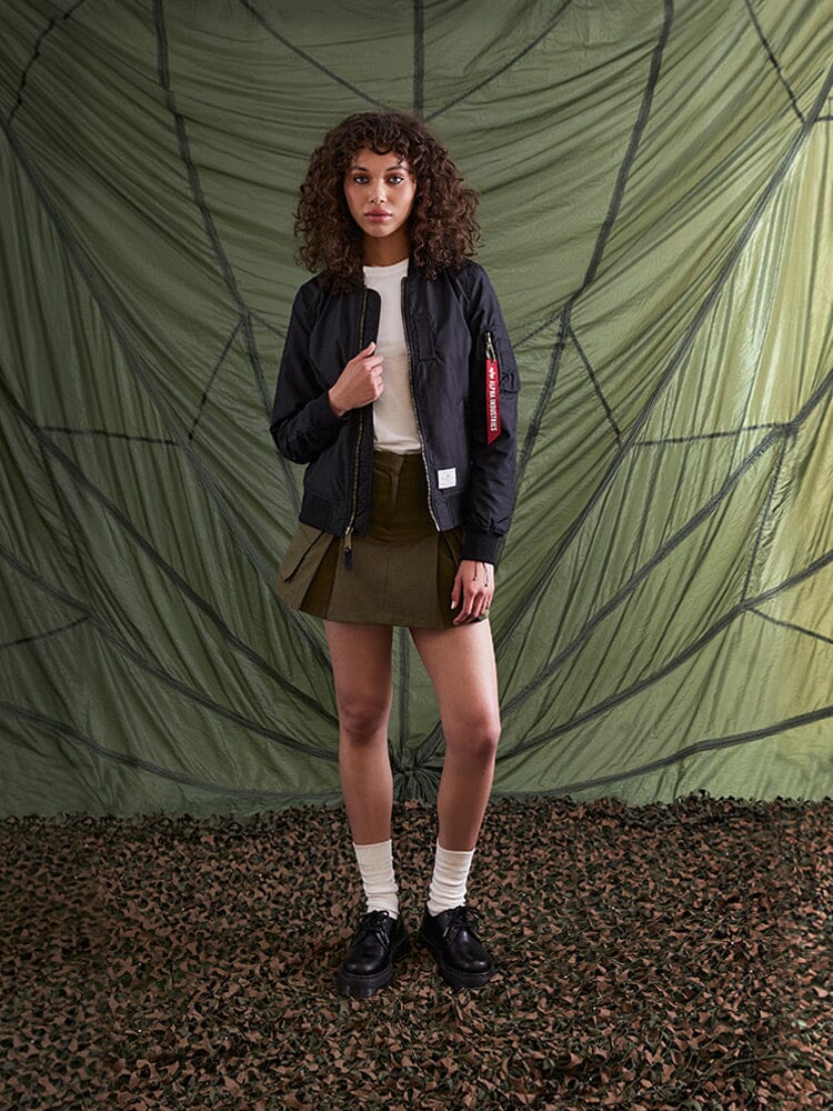 A person with curly hair stands wearing the Alpha Industries L-2B Skymaster Gen II Bomber Jacket, along with a white shirt, olive skirt, and black shoes paired with white socks.