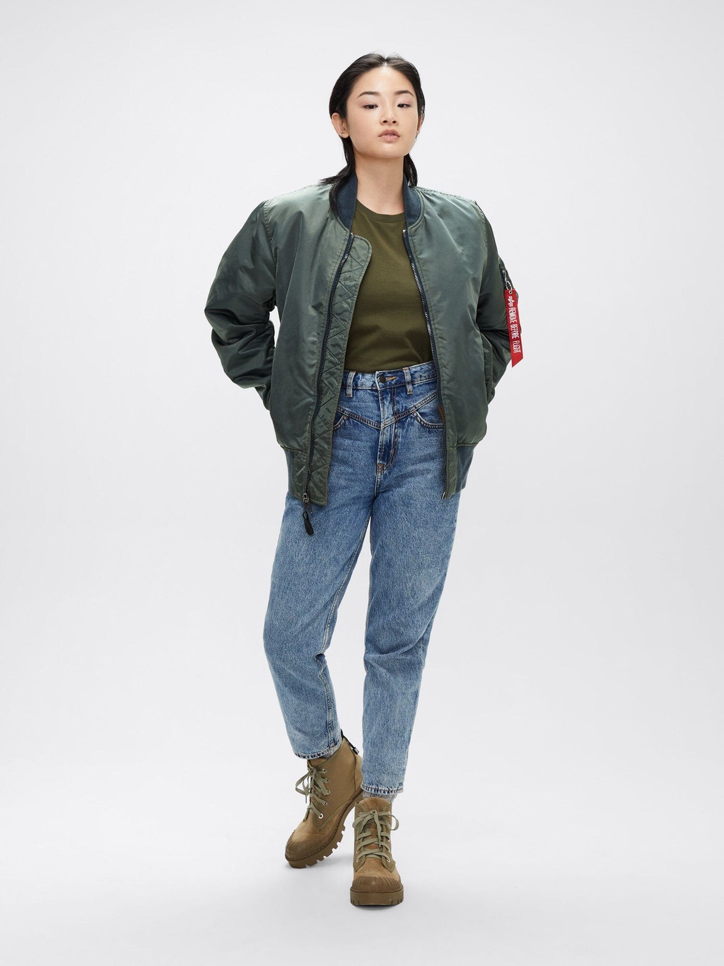 A person wearing a green MA-1 Battlewash Bomber Jacket by Alpha Industries, Inc., paired with a green shirt, blue jeans, and brown boots.
