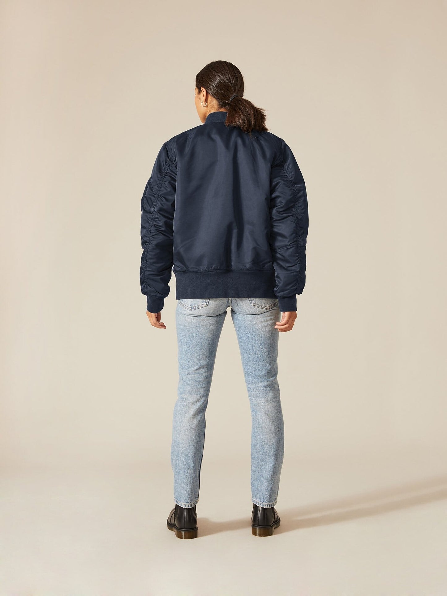A person wearing an Alpha Industries MA-1 Battlewash Bomber Jacket in navy, accompanied by light blue jeans and black shoes, stands with their back to the camera.